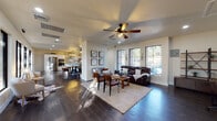 This well-appointed single-story home offers a seamless open-concept layout that connects the living, dining, and kitchen areas into one cohesive, flowing space. Dark hardwood floors run throughout, providing a striking contrast against the crisp white walls and trim.

The living area is generously proportioned, bathed in natural light streaming through a series of large black-framed windows that line the exterior wall. A ceiling fan, recessed lighting, and wall sconces contribute to a comfortable and well-lit atmosphere, while the layout accommodates both relaxed seating and a dedicated workspace area.

The dining space centers around a substantial farmhouse-style table and is anchored by a floor-to-ceiling stone fireplace with a mounted TV above — a standout architectural feature that adds warmth and character to the room. Additional black-framed windows frame outdoor views on multiple sides.

The kitchen is a highlight of the home, featuring white shaker-style cabinetry with matte black hardware, light quartz countertops, a subway tile backsplash, a built-in wine rack, and a central island with pendant lighting overhead. Stainless steel appliances — including a double wall oven and French door refrigerator — complete this highly functional culinary space.

A long hallway connects the main living areas to the rest of the home, lined with additional windows that ensure natural light reaches every corner of this thoughtfully designed residence.