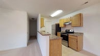 This single-floor, three-bedroom space offers a practical and well-organized layout designed for comfortable everyday living. The kitchen serves as a central feature, showcasing warm honey-toned wood cabinetry that provides generous storage both above and below the countertops. Dark granite-style countertops contrast nicely with the lighter cabinetry, adding a touch of visual interest. Stainless steel appliances, including a refrigerator and an electric range with a built-in overhead vent hood, give the kitchen a modern, functional feel. The tile flooring in the kitchen is easy to maintain and transitions naturally into the adjoining areas. A peninsula-style counter extends outward, creating a natural divide between the kitchen and the adjacent carpeted living area, offering potential for casual dining or additional prep space. A hallway extending from the kitchen area leads to the remainder of the unit, connecting the living spaces to the three bedrooms. The carpeted hallway is well-lit with ceiling-mounted fixtures, contributing to a clean and welcoming atmosphere throughout. Overall, the space presents a cohesive, move-in-ready environment with a straightforward and efficient floor plan.