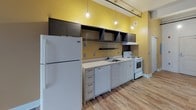 This single-floor space offers an open, airy layout characterized by clean white walls and warm wood-look flooring that extends throughout the main living area. A row of tall, black-framed windows lines one wall, allowing generous natural light to fill the expansive open floor plan while providing urban rooftop views. An industrial-style track rail runs along the ceiling, adding a contemporary, loft-like aesthetic to the space.

The kitchen is a standout feature, set against a vibrant yellow accent wall that lends a bold, energetic character. It is fully equipped with white cabinetry, open dark-toned upper shelving, a white electric range, dishwasher, refrigerator, and a single-basin sink with light countertops. Pendant bulb lighting overhead adds a warm, functional ambiance to the cooking area. A wall-mounted mini-split HVAC unit ensures climate comfort throughout the space.

The bathroom is cleanly appointed with a wall-mounted sink, a rectangular mirror with overhead lighting, and a tiled shower stall. An adjacent open closet area with a hanging rod provides convenient storage. Overall, the space combines industrial design elements with practical finishes, creating a functional and visually engaging environment suitable for a variety of uses.