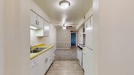 This single-floor unit offers a practical and straightforward layout designed for comfortable everyday living. The kitchen is equipped with dark wood cabinetry — both upper and lower — paired with a light butcher-block-style countertop, a double-basin stainless steel sink, an electric range, and a full-size refrigerator. Stone-pattern vinyl tile flooring runs through the kitchen and connects to the adjacent living area, creating a cohesive flow between spaces. The living area is generously sized, carpeted, and includes a wall-mounted air conditioning unit alongside baseboard heating, ensuring year-round climate control. The bedrooms are carpeted and bright, each featuring windows fitted with horizontal blinds that allow natural light to filter in, along with baseboard heating for added comfort. The bathroom includes a combination tub and shower, a toilet, a vanity with mirror, and warm wood-tone flooring that adds a touch of character. Throughout the unit, neutral wall tones and clean white trim create a simple, understated aesthetic. The overall layout, as seen in the dollhouse view, is well-organized and efficiently utilizes the available space across its single level.