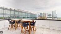This contemporary urban space combines a well-appointed indoor kitchen with an expansive outdoor rooftop terrace, offering a seamless indoor-outdoor experience with striking city skyline views. The rooftop terrace is finished with large-format porcelain tile flooring and features multiple high-top wooden dining tables paired with modern dark chairs, providing ample seating for gatherings. A stainless steel gas grill adds functionality for outdoor entertaining, while a low glass perimeter barrier ensures unobstructed panoramic views of the surrounding cityscape and active urban development. Corrugated metal cladding along the exterior walls gives the terrace a sleek, industrial character. Floor-to-ceiling glass sliding doors connect the terrace directly to the interior, flooding the space with natural light and maintaining a visual connection to the outdoors. The kitchen is thoughtfully designed with white flat-front cabinetry, dark countertops, and a vibrant blue vertical tile backsplash that serves as a bold focal point. Stainless steel appliances, including a gas range and built-in microwave, are complemented by a central island and polished concrete flooring. Angular windows frame additional city views, reinforcing the elevated urban setting throughout.
