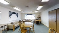 This single-floor space features an open, combined living and dining layout designed for comfortable everyday use. The room is finished with a textured ceiling and illuminated by several recessed fluorescent light panels, providing consistent, even lighting throughout. A central ceiling vent contributes to climate control across the space.

The flooring is covered in soft, light blue carpeting, lending a relaxed and welcoming atmosphere to the room. Two dining tables with wooden Windsor-style chairs are positioned centrally, offering ample seating for multiple occupants. Along the left wall, a striped upholstered sofa provides additional casual seating, complemented by a loveseat positioned near the window.

Natural light enters through a set of windows dressed with dark navy valances and curtains, adding a decorative touch to the otherwise neutral color palette of white and beige walls. Near the windows, a decorative side table holds a floral arrangement, adding a homey accent to the space. A wooden hutch or buffet piece lines the far right wall, offering functional storage.

Overall, the space presents a modest, functional, and comfortable communal environment suitable for dining, relaxing, and gathering.