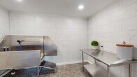 This single-floor pet grooming facility combines functional design with refined finishes to create a clean, professional environment. The space is organized around a central hallway finished with hexagonal tile flooring in a neutral gray tone, with multiple doors providing access to individual grooming rooms. A wall-mounted mini-split system ensures comfortable climate control throughout.

The grooming rooms are uniformly finished with large-format, marble-look ceramic wall tiles in white with subtle gray veining, lending an upscale character to the otherwise utilitarian space. Recessed lighting provides consistent, even illumination in each room. The flooring throughout the grooming areas continues the hexagonal tile pattern, offering both durability and visual cohesion.

Each grooming room is equipped with stainless steel fixtures, including a professional-grade elevated bathing tub with an attached hose and spray nozzle, as well as stainless steel work tables with under-shelf storage. These surfaces are easy to clean and built for heavy daily use. The facility concludes with an exterior-facing door at the end of the hallway, allowing natural light to filter into the space.