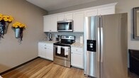 Located on E Coliseum Blvd in Fort Wayne, Indiana, this single-floor unit presents a well-designed layout with a consistent contemporary aesthetic carried throughout each room. Warm taupe and gray tones form the color palette, accented by bold yellow details that add energy and visual interest across the space.

The living room is a standout area, featuring an impressive bank of tall windows fitted with white horizontal blinds that flood the room with natural light and offer outdoor views of the surrounding area. Plush carpeting and a generous sectional sofa create a comfortable, welcoming atmosphere.

Adjacent to the living room, the dining area continues the window-rich design, with additional tall windows providing ample daylight. The space comfortably accommodates a round dining table, making it well-suited for entertaining.

The kitchen is outfitted with white shaker-style cabinetry, granite-look countertops, and a full suite of stainless steel appliances, including a range, over-the-range microwave, and French door refrigerator. Wood-look flooring anchors the space with a clean, modern feel.

Two bedrooms offer carpeted floors and natural light through blinds-covered windows. The bathroom includes a tub-shower combination with wood-look flooring. Overall, the unit maintains a cohesive, polished aesthetic throughout.