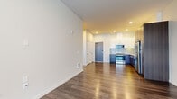 This well-designed one-bedroom, one-bathroom unit offers a thoughtfully arranged single-floor layout that maximizes functionality within a compact footprint. The open-concept living and kitchen area forms the heart of the unit, anchored by rich hardwood-style flooring that extends throughout the main living spaces. The contemporary kitchen is a standout feature, showcasing two-tone cabinetry—glossy white upper cabinets paired with dark wood-finish lower cabinets—complemented by a light subway tile backsplash, quartz countertops, and a full suite of stainless steel appliances including a range, microwave, dishwasher, and refrigerator. Recessed lighting illuminates the kitchen and entry area effectively. The living room provides an open, versatile space with clean white walls and consistent hardwood-style flooring. The bedroom is a comfortable, well-proportioned room featuring plush carpet, a large window that admits natural light, and a clean, neutral palette. The bathroom continues the modern aesthetic with wood-tone cabinetry, a white quartz vanity top, a large mirror, and a full tub-shower combination with tile surround and hardwood-style flooring.