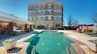Located at 5437 Bowman Rd in Macon, Georgia, this outdoor amenity area is defined by its classic brick architecture and well-appointed leisure spaces. The centerpiece is a generously sized, freeform swimming pool with clear blue-green water, complemented by an adjacent spa. The pool deck is finished with attractive paver stonework and is surrounded by lounge chairs and shaded umbrella seating, offering ample space for relaxation under the open sky. The multi-story brick building backdrop, with its large grid-pane windows, frames the outdoor area and provides a refined architectural context. A covered outdoor pavilion serves as a true gathering space, featuring exposed brick columns, a coffered ceiling with a ceiling fan, and a full brick fireplace equipped with a mounted flat-screen television — ideal for year-round outdoor entertaining. Adjacent seating includes both a dining table with chairs and a plush sectional sofa, creating a comfortable and versatile lounge environment. The pavilion opens directly toward the pool area, allowing for seamless indoor-outdoor flow. Lush, manicured shrubs and potted plantings add greenery throughout the space, enhancing the welcoming atmosphere of this well-designed outdoor retreat.