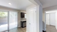 This single-floor residence offers a clean, contemporary design with a cohesive color palette of soft grays and whites that creates a calm and inviting atmosphere throughout. The kitchen stands out as a focal point, showcasing rich dark cabinetry paired with light stone-look countertops and a distinctive mosaic tile backsplash that adds texture and visual interest. A stainless steel dishwasher adds a modern, functional touch to the space. The kitchen benefits from natural light streaming through a window, keeping the area bright and airy. A sliding glass door in the adjacent living area provides additional natural light and outdoor access, fitted with vertical blinds for privacy and light control. Durable wood-look flooring runs throughout the main living areas, offering both style and practicality. Crisp white panel doors with clean trim work are featured throughout, contributing to the home's fresh, updated aesthetic. The layout flows naturally from the main living and kitchen area into a hallway leading to additional rooms, providing a functional and well-organized floor plan. Overall, the space presents a modern, move-in-ready feel with thoughtful finishes and a neutral design suited to a variety of styles.
