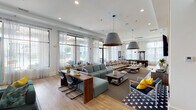 This thoughtfully designed single-floor amenity space blends social, recreational, and professional functions into one cohesive environment. The expansive main lounge serves as the centerpiece, featuring soaring ceilings, large floor-to-ceiling windows dressed with sheer curtains, and an abundance of natural light. Eclectic seating arrangements — including a bold plaid sectional, a teal modular sofa, and accent chairs with vibrant yellow cushions — are anchored by striking oversized pendant lights. A wood-paneled feature wall with a mounted flat-screen TV and built-in credenza adds a refined focal point, while a pool table in the background reinforces the space's recreational appeal. Light hardwood flooring and patterned area rugs add warmth and texture throughout. A dedicated business center offers computer workstations, built-in cabinetry, and shelving beneath clerestory windows. Multiple meeting rooms provide intimate settings for focused work, each outfitted with conference tables, sleek seating, wall-mounted displays, and decorative artwork. A cozy nook with bench seating and deep green accent walls offers a quiet retreat. Throughout, curated artwork and a carefully layered mix of materials create a vibrant yet functional atmosphere suited to a variety of needs.
