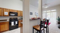 This thoughtfully designed single-floor unit offers a cohesive layout that maximizes comfort and functionality. The living area is an immediate standout, boasting dramatically tall ceilings with crown molding, plush carpeting, and a ceiling fan. Floor-to-ceiling sliding glass doors flood the space with natural light and provide access to an outdoor area, while transom windows above amplify the airy, open atmosphere.

The kitchen is well-appointed with warm honey-toned wood cabinetry, dark granite-style countertops, tile flooring, and a full suite of appliances including a stainless steel refrigerator, black electric range, and built-in microwave. A peninsula with a breakfast bar seamlessly connects the kitchen to the dining and living areas, making it ideal for casual dining and entertaining.

The bedrooms are carpeted and feature impressively tall ceilings that enhance the sense of space. The primary bedroom includes direct access to a private bathroom, while ample closet space is provided throughout. Neutral wall tones carry through the entire unit, creating a clean and versatile backdrop. Overall, the space offers a bright, open ambiance with a practical and well-considered design.