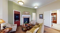 Located at 400 E Glade Rd in Grapevine, Texas, this single-story community facility offers a thoughtfully designed layout with a variety of functional and comfortable common spaces. The inviting lounge serves as a central gathering area, featuring a decorative fireplace with a stone surround, a built-in media cabinet in rich cherry wood, crown molding, and warm sage green accent walls complemented by neutral tones. Comfortable seating arrangements and elegant pendant lighting create a welcoming ambiance throughout. The formal dining room is bright and spacious, benefiting from an abundance of natural light through large windows. It accommodates generous seating around a long wooden dining table, with additional cabinetry and counter space along one wall. An adjoining casual dining area offers direct outdoor access through a glass-paned door and includes a small kitchenette with built-in cabinetry and a microwave. The library is well-appointed with floor-to-ceiling cherry wood bookshelves stocked with a wide collection of books, carpeted flooring, and soft overhead lighting for a quiet, studious atmosphere. A dedicated workstation nook features built-in cherry cabinetry and counter space for computing and media needs. The fitness room is equipped with cardio machines including a stationary bike and elliptical, and is well-lit by multiple windows with neighborhood views and a ceiling fan for ventilation.