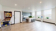 Located at 74 Franklin St in downtown Buffalo, NY, this single-floor communal amenity space offers a well-appointed, open-plan layout designed for both relaxation and social interaction. The space is finished with light hardwood flooring throughout, crisp white ceilings with recessed lighting, and clean white walls that contribute to a bright, airy atmosphere.

The kitchen area is a standout feature, equipped with sleek grey cabinetry, white subway tile backsplash, stainless steel appliances, and a large center island with a butcher block countertop and bar seating—ideal for casual gatherings. A water cooler and accessible restroom are conveniently located nearby.

The lounge area is generously sized, anchored by a large sectional leather sofa, an upholstered ottoman, and a patterned area rug, all positioned beneath large windows that flood the room with natural light and offer views of the surrounding Buffalo cityscape. A wall-mounted flat-screen TV and a pair of accent chairs create a secondary seating zone.

A games area features a full-length shuffleboard table set against an exposed brick wall adorned with framed architectural prints, adding a warm, industrial character to the space. Potted plants are distributed throughout, enhancing the welcoming ambiance of this versatile communal retreat.