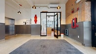 Located at 450 Amsterdam St in Detroit's vibrant Midtown neighborhood, this single-floor commercial space masterfully merges industrial character with contemporary design. The welcoming entry lobby features polished terrazzo floors, exposed brick walls, pendant lighting with copper-toned fixtures, and a sleek reception area framed by black steel-and-glass partitions. An elevator and staircase provide internal access, while large glass block windows flood the space with natural light.

Beyond the lobby, a spacious lounge area showcases polished red concrete floors, whitewashed brick walls, and oversized black-framed industrial windows that offer views of the surrounding cityscape. A warm leather sofa and modern chandelier anchor this inviting gathering space. Adjacent to it, a multi-use room serves as both a conference and collaborative area, equipped with a wall-mounted display, a central table, and high-top seating along the window line.

A well-designed kitchenette features light wood cabinetry, white quartz countertops, a subway tile backsplash, open shelving, and stainless steel appliances. A long corridor lined with dark paneling connects the various zones, while a mailroom area with built-in cubbies and large windows completes the layout. Throughout, exposed ductwork and high ceilings reinforce the space's industrial-modern aesthetic.