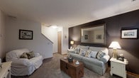 Located at 1600 Belvoir Boulevard in Columbus, Ohio, this two-floor residence offers a well-organized and comfortable layout spread across two levels. The main living area is a welcoming space anchored by plush wall-to-wall carpeting that adds warmth underfoot. A striking dark accent wall serves as a focal point, adorned with a large decorative mirror that reflects light and enhances the sense of depth in the room. The living space accommodates ample seating and features a rustic wooden trunk coffee table that adds a charming, eclectic touch to the overall aesthetic. Neutral white walls throughout the main level create a clean and versatile backdrop, while ceiling lighting provides consistent illumination. A staircase situated along the interior wall leads to the upper floor, offering a clear and accessible transition between levels. An interior door near the base of the staircase provides access to additional utility or storage space. The overall ambiance of the home is cozy and inviting, blending neutral tones with thoughtful design accents. The two-story layout provides a natural separation between living and private spaces, making this Columbus residence a functional and comfortable home.