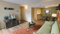 This single-floor residential space offers a well-connected open layout that integrates the living, dining, and kitchen areas into one cohesive environment. The living area features rich, dark hardwood-style flooring that extends throughout the space, anchored by a warm, textured area rug that adds depth and color contrast. A round coffee table serves as the centerpiece of the seating area, complementing the relaxed, contemporary feel of the room.

A distinct accent wall painted in a deep green adds a bold design element to the dining area, which comfortably accommodates a table and chairs. The dining space flows naturally into the kitchen, which is partially visible through a pass-through window. The kitchen features light countertops and a clean, functional layout, providing an efficient workspace while maintaining visual openness with the adjacent rooms.

The neutral beige tones of the walls throughout the main living areas create a warm and inviting backdrop, while visible ceiling vents indicate a central HVAC system for year-round comfort. A hallway leads to additional rooms, suggesting a practical and well-organized floor plan. Overall, the space presents a balanced combination of comfort, functionality, and contemporary design details suited to everyday living.