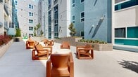 Located at 1642 E Florence Ave in Los Angeles, CA, this modern residential building presents a striking multi-story facade finished in a contemporary palette of blue and grey tones, accented by teal-framed windows and balconies. The architecture is clean and geometric, with well-defined horizontal lines that give the structure a bold, urban character.

At the heart of the building lies a spacious open-air courtyard that serves as a central gathering space for residents. The courtyard is thoughtfully designed with multiple seating zones, including a lounge area furnished with vibrant orange modular chairs arranged around a round side table, as well as casual dining seating with light grey chairs and white tables. Overhead string lights add a warm, welcoming ambiance ideal for evening gatherings.

Raised masonry planters constructed of textured concrete block are distributed throughout the courtyard, featuring a curated mix of drought-tolerant plants, succulents, ornamental grasses, and shade trees that bring natural greenery to the space. A dedicated children's play area is also integrated into the courtyard, featuring colorful play structures enclosed within its own section of the outdoor space.

Wide, smooth concrete pathways connect the various areas of the courtyard, providing accessibility throughout. Unit entry doors, accented in teal and blue, open directly onto the ground-level walkway, creating a seamless indoor-outdoor connection for residents.
