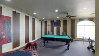 This single-story, 2-bedroom, 2-bathroom home includes a standout game room designed for entertaining. The game room is a spacious, carpeted area anchored by a full-size pool table with a green felt surface and ornate dark wood legs. A wall-mounted flat-screen TV is positioned prominently on one wall, making the space equally suited for viewing and recreation. Pool cues are neatly stored on a rack nearby, and additional seating, including a curved chair with built-in cup holders, adds a lounge-like atmosphere. The walls feature bold vertical brown and cream stripes, giving the room a dynamic, stylized look. Recessed lighting provides bright, even illumination throughout, while a ceiling fan offers comfort and air circulation. The textured ceiling and clean white trim provide a polished contrast to the bold wall design. The overall ambiance of this room is energetic and well-appointed, making it a functional and visually engaging recreational space within the home.