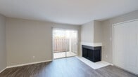 This single-floor residence offers a clean, neutral aesthetic with wood-look flooring running throughout the main living areas. The living room serves as the focal point of the space, showcasing a corner fireplace with a white surround and dark insert, adding both visual interest and functional warmth. A full-width sliding glass door fitted with vertical blinds floods the room with natural light and opens onto a private, fenced patio, providing a seamless indoor-outdoor connection.

An additional open room adjacent to the living area features the same wood-look flooring and neutral wall tones, offering flexible use as a dining area or secondary living space. The kitchen is outfitted with white cabinetry, white countertops, a subway tile backsplash, and a full suite of stainless steel appliances, including an electric range, over-the-range microwave, and refrigerator.

The bedroom features wood-look flooring, a ceiling fan with light fixture, a window with exterior views, and a textured ceiling. The bathroom includes a full tub-shower combination with white tile surround, wood-look tile flooring, and clean white fixtures. Throughout the unit, consistent neutral tones and updated finishes contribute to a cohesive, move-in ready atmosphere.