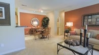 This single-story residence features an open-concept layout that connects the living and dining areas into one cohesive, functional space. The interior is defined by striking terracotta-toned accent walls that add warmth and visual depth, contrasting elegantly against the neutral cream-colored base walls and light carpeted flooring throughout.

The dining area accommodates a round wooden table with matching cross-back chairs, complemented by a decorative sunburst mirror and a tall indoor plant that adds a natural, organic element to the space. A sideboard beneath the mirror provides additional surface space and storage.

The living area features a plush, neutral-toned sofa accented with warm-hued throw pillows, positioned alongside a glass-top coffee table with a dark metal frame. A table lamp with a classic white shade provides ambient lighting near the seating area.

Track lighting mounted along the ceiling supplements the overall illumination and adds a modern touch. Crown molding along the ceiling edges refines the space with a polished, finished look. A doorway visible in the background hints at additional rooms beyond, suggesting a well-organized and thoughtfully designed floor plan throughout.