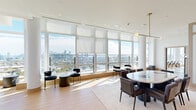 Located at 54 Jones St in Jersey City, NJ, this elevated coworking space offers a refined, modern environment designed for productivity and collaboration. Floor-to-ceiling windows wrap the perimeter, flooding the interior with abundant natural light and framing sweeping panoramic views of the Jersey City and Manhattan skylines, as well as the surrounding waterways.

The dining area is a standout feature, anchored by a striking marble-topped communal table surrounded by contemporary seating. Elegant brass pendant lighting hangs overhead, complementing the white tile flooring with navy accent inlays. Smaller bistro-style tables along the window line provide additional seating options with unobstructed views.

The living room area offers a more relaxed setting, furnished with a plush neutral sofa, a veined black marble coffee table, and curated artwork. A bold brass dome pendant light adds warmth to the space, while corner floor-to-ceiling glass panels maximize the breathtaking skyline backdrop.

Semi-private booth seating areas, defined by textured partition screens and topped with brass pendant lights and marble-surfaced tables, provide intimate nooks for focused work or small meetings. Light wood flooring runs through these zones, lending warmth and continuity. A hallway connects the areas, completing this thoughtfully designed, light-filled coworking environment.