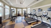 This fitness center is designed with both functionality and architectural elegance in mind. The space is anchored by a striking octagonal rotunda with a coffered, tray-style ceiling accented by crown molding and a ceiling fan, creating a sense of openness and volume overhead. Recessed lighting throughout ensures the space is well-lit at all times.

The flooring features a bold checkerboard pattern in warm tan and deep brown tones, adding visual interest across the entire workout area. A wraparound bank of large windows in the rotunda section floods the space with natural light and provides views of the surrounding outdoor environment, enhancing the overall workout atmosphere.

The cardio zone is well-equipped with multiple True-brand treadmills and elliptical machines, each outfitted with personal display screens. Wall-mounted televisions are positioned for easy viewing during workouts. The strength training area includes a multi-functional cable machine, a weight bench, dumbbells, and various resistance equipment arranged to maximize usable floor space.

Glass-paneled French doors serve as the main entry, lending a refined, welcoming aesthetic. The sage green walls, white millwork, and thoughtful layout combine to create a polished, comfortable environment suited for a comprehensive fitness routine.