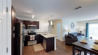 This single-floor space showcases an open-concept layout that seamlessly connects the kitchen, dining, and living areas. The kitchen is a standout feature, equipped with stainless steel appliances including a refrigerator, range, microwave, and dishwasher. Rich, dark espresso cabinetry provides ample storage and contrasts beautifully with the light granite-style countertops. A peninsula with an undermount sink serves as both a functional workspace and a natural divider between the kitchen and living areas. Three sleek pendant lights hang above the peninsula, adding a modern, polished touch to the space. Warm hardwood-style flooring flows throughout, lending continuity and a sense of warmth to the entire unit. The living area is bright and welcoming, with neutral walls that offer versatility for a variety of décor styles. Recessed and track lighting complement the pendant fixtures, ensuring the space is well-lit throughout. A hallway visible from the main area suggests additional rooms beyond, contributing to the overall functional flow of the layout.