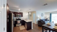 This single-floor space showcases an open-concept layout that seamlessly connects the kitchen, dining, and living areas. The kitchen is a standout feature, equipped with stainless steel appliances including a refrigerator, range, microwave, and dishwasher. Rich, dark espresso cabinetry provides ample storage and contrasts beautifully with the light granite-style countertops. A peninsula with an undermount sink serves as both a functional workspace and a natural divider between the kitchen and living areas. Three sleek pendant lights hang above the peninsula, adding a modern, polished touch to the space. Warm hardwood-style flooring flows throughout, lending continuity and a sense of warmth to the entire unit. The living area is bright and welcoming, with neutral walls that offer versatility for a variety of décor styles. Recessed and track lighting complement the pendant fixtures, ensuring the space is well-lit throughout. A hallway visible from the main area suggests additional rooms beyond, contributing to the overall functional flow of the layout.