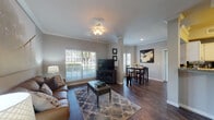 This single-floor residence offers a thoughtfully designed layout with a seamless flow between its primary living spaces. The living room serves as the heart of the home, featuring dark hardwood-style flooring, crown molding, and large windows fitted with blinds that invite generous natural light and outdoor views. A decorative column subtly defines the transition into the dining area, which accommodates a bistro-height table and chairs beneath recessed lighting.

The kitchen is outfitted with crisp white cabinetry, granite-style countertops, a mosaic tile backsplash, and a full suite of black appliances including a range, microwave, dishwasher, and refrigerator. The hardwood-style flooring continues here, maintaining a consistent aesthetic.

Two bedrooms provide comfortable retreats, both carpeted for warmth. The primary bedroom features vaulted ceilings, a walk-in closet, and sliding glass doors opening to a private outdoor patio with greenery views. The secondary bedroom is quietly tucked away with its own closet access.

The bathroom presents a clean, functional design with granite-style vanity countertops, white cabinetry, a tub-shower combination with tile surround, and hardwood-style flooring continuing through the space.