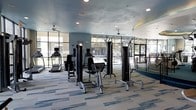 This fitness center offers a bright, contemporary workout environment designed to support a full range of exercise routines. The space is anchored by a selection of strength training machines and cardio equipment, including treadmills and multi-functional cable and resistance machines, all arranged to allow comfortable movement between stations. Large floor-to-ceiling windows line the perimeter, providing abundant natural light and open views of the surrounding outdoor landscape, creating an inviting and energizing atmosphere. The ceiling features recessed lighting alongside ceiling fans, ensuring the space remains well-lit and well-ventilated during use. The flooring consists of patterned carpet tiles in blue and grey tones, adding a modern aesthetic while providing comfort underfoot. The walls are finished in a soft blue-grey palette that complements the overall contemporary design. The generous layout allows ample space between equipment, promoting ease of use and an uncluttered workout experience. With its combination of functional equipment, thoughtful design, and plentiful natural light, this fitness center provides a welcoming and professional environment suited for both focused strength training and cardiovascular exercise.