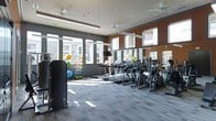 This contemporary fitness center is designed to provide a comprehensive workout experience in a bright, welcoming environment. Large floor-to-ceiling windows line the perimeter of the space, flooding the interior with natural light and offering views of the surrounding exterior. The open layout accommodates a generous selection of cardio equipment, including treadmills and elliptical trainers, arranged in orderly rows to maximize usability and flow. Ceiling fans are strategically positioned throughout, ensuring consistent air circulation during workouts. Full-length mirrors are mounted along the walls, enhancing the sense of space while allowing members to monitor their form. A dedicated functional training and stretching zone is visible in the corner, stocked with stability balls, resistance equipment, and exercise mats in vibrant colors, adding energy to the room. The interior design features a bold color palette of warm orange and charcoal gray accent walls, complemented by durable, textured flooring suitable for high-traffic athletic use. Overhead lighting fixtures supplement the natural light for consistent illumination at any time of day. The overall ambiance is clean, motivating, and professionally appointed, making it a well-rounded facility for a variety of fitness routines.