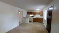 This single-floor space offers a practical and functional layout that combines the living area and kitchen in an open-concept design. The kitchen is well-equipped with warm medium-toned wood cabinetry providing ample upper and lower storage, white appliances including a range, refrigerator, and a double-basin sink positioned beneath a bright window that allows natural light to filter in. A kitchen peninsula offers a convenient separation between the cooking and living spaces while maintaining an open flow. The flooring transitions from tile in the kitchen area to carpet in the living space, adding a sense of defined zones within the open layout. A baseboard heater along the exterior wall provides heating to the space. The textured ceiling adds a classic touch throughout. A doorway visible from the main living area leads to additional rooms, suggesting a connected and accessible floor plan. The overall ambiance is clean, bright, and functional, making it a straightforward and livable space suited for everyday use.