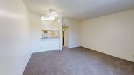 This single-floor space features an open and functional layout designed for comfortable everyday living. The main living area is finished with wall-to-wall carpeting in a neutral gray tone, providing a soft, cohesive foundation throughout the room. Cream-colored walls keep the space feeling bright and airy, while a ceiling fan with a built-in light fixture serves as both a practical and central overhead element.

A half-wall pass-through connects the living area to the kitchen, creating an open sightline between the two spaces while maintaining a degree of separation. The kitchen is equipped with standard appliances, including a refrigerator and range with an overhead ventilation hood, and features a countertop that extends along the pass-through, offering casual dining or prep space. Cabinetry above provides additional storage. A hallway visible from the living area leads further into the unit, suggesting additional rooms beyond. The textured ceiling adds a subtle architectural detail consistent throughout the space. Overall, the layout is straightforward and well-organized, making efficient use of the available floor plan.