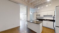 Located at 24 Mt Vernon St in Lynn, MA, Unit 201 is a striking loft-style unit that blends industrial character with modern finishes. The space is defined by soaring ceilings with exposed structural beams painted white, polished concrete floors, and sturdy support columns that reinforce its converted-building aesthetic. Track lighting runs throughout, illuminating the open layout with a bright, airy ambiance.

The kitchen is a standout feature, showcasing an abundance of white cabinetry, dark granite countertops, and a central island with a matching granite surface. Stainless steel appliances — including a refrigerator, range, and microwave — complete the well-appointed culinary space. Pendant lighting above the island adds a refined touch to the otherwise industrial character.

The open living area extends beyond the kitchen, offering generous, flexible space accentuated by white walls and polished concrete underfoot. A barn-style sliding door adds an architectural detail that echoes the loft's industrial roots. Natural light filters in through large windows, enhancing the sense of openness throughout.

The bathroom is finished with large-format dark slate-look floor tiles, white subway tile in the tub surround, and clean white cabinetry, offering a crisp, contemporary contrast to the raw industrial elements found elsewhere in the unit.