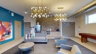 This single-floor space is a thoughtfully designed communal lounge that seamlessly blends modern aesthetics with functional amenities. The room makes an immediate visual impact with its pair of dramatic gold branch-style chandeliers, whose exposed globe bulbs cast a warm, inviting glow across the entire area. A textured stacked-stone accent wall serves as the architectural centerpiece, anchoring the built-in kitchenette that is equipped with a stainless steel refrigerator, microwave, sink, and a coffee station — providing all the essentials for a comfortable communal experience. The lounge area is furnished with a contemporary gray sofa, matching accent chairs, and a low-profile coffee table, all arranged on a plush area rug to define the seating zone. A vibrant teal accent wall adds a bold pop of color, contributing to the space's energetic yet relaxed atmosphere. Exposed ductwork on the ceiling lends an industrial character that contrasts pleasingly with the refined furnishings below. The open layout and generous ceiling height create a sense of spaciousness, making this lounge well-suited for both quiet relaxation and social interaction.