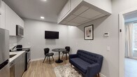 This well-designed single-floor space showcases a clean, contemporary aesthetic carried consistently throughout every room. Light-toned hardwood flooring runs seamlessly across the unit, complementing the neutral gray and white walls that create a bright, cohesive atmosphere.

The kitchen is a standout feature, outfitted with two-tone gray cabinetry — darker lower cabinets paired with lighter upper ones — along with white quartz countertops, a white square-tile backsplash with under-cabinet lighting, a stainless steel sink, and stainless steel appliances including a range and over-the-range microwave. A peninsula layout maximizes functionality and flow.

The lounge area provides a comfortable gathering space, furnished with a tufted navy sofa, a round bistro table with modern chairs, a wall-mounted flat-screen TV, and framed wall art. Recessed lighting and acoustic ceiling panels contribute to the room's refined, functional character.

The bedroom is generously proportioned, featuring multiple windows dressed with light linen curtains that allow natural light to filter in, recessed ceiling lights, and ample open floor space.

The bathroom is finished to a high standard, featuring a large walk-in glass-enclosed shower with matte black hardware and fixtures, a floating wood-toned vanity, and dark tile flooring.

A clean, well-lit hallway with white panel doors and matte black hardware connects the rooms, reinforcing the space's modern and polished design language.