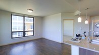 This single-floor space offers a clean, modern design characterized by an open-concept layout that seamlessly connects the living area to the rest of the unit. Dark hardwood flooring runs throughout, providing a rich contrast against the light gray walls and white ceiling, creating a contemporary and inviting atmosphere.

The living area is brightened by a large picture window that frames views of the surrounding urban environment, allowing natural light to fill the space. A flush-mount ceiling fixture provides additional overhead lighting. The neutral wall tones and simple finishes give the space a versatile, move-in-ready appeal.

A partial view of the kitchen area is visible in the foreground, featuring a granite countertop surface that extends as a breakfast bar-style divide between the kitchen and living space. A doorway with light wood trim leads into an adjoining room, suggesting a functional and well-connected floor plan. The overall ambiance is understated and modern, with clean lines and practical finishes throughout.