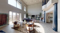 This impressive two-story space is defined by its dramatic architectural design and open, airy atmosphere. The centerpiece is a grand great room boasting soaring double-height ceilings lined with arched windows that flood the interior with natural light. Light-toned tile flooring and recessed lighting enhance the sense of openness, while French doors provide direct access to an outdoor balcony with views of the surrounding neighborhood. The living area is thoughtfully arranged with a plush seating arrangement, glass-top coffee tables, and a large decorative wood-panel wall accent that adds warmth and character to the space.

An ornate wrought-iron balcony railing on the second floor overlooks the great room below, connecting the two levels with visual elegance. The upper level houses a dedicated game room complete with a full-sized billiards table, a wall-mounted television, and a poker table seating area — ideal for recreation and entertainment.

The kitchen is clean and functional, featuring crisp white shaker cabinetry, sage-green subway tile backsplash, white quartz countertops, and stainless steel appliances including a side-by-side refrigerator and microwave. Wood-look tile flooring ties the kitchen seamlessly to the rest of the space. Throughout, the design balances modern finishes with classic architectural details, creating a polished and inviting environment.