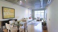 Model #733 presents a thoughtfully designed single-floor space that seamlessly blends industrial and contemporary aesthetics. The exposed concrete ceiling serves as a defining architectural feature, complemented by visible ductwork and track lighting that lend the space an authentic loft character. Hardwood flooring runs throughout, adding warmth that balances the raw industrial elements overhead.

The open-plan layout integrates the dining and living areas into one cohesive, flowing space. The dining area accommodates a glass-top table with upholstered seating, while the living area is anchored by a comfortable seating arrangement centered around a glass coffee table atop a area rug. The two zones coexist naturally, creating an inviting, functional environment.

Large gridded windows along the far wall flood the interior with natural light and offer outward views of the surrounding urban landscape, enhancing the sense of openness. An accent wall in a muted sage green adds a subtle pop of color, softening the otherwise neutral palette of whites and light tones. Decorative greenery throughout the space adds life and texture, contributing to the overall welcoming ambiance.