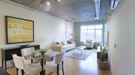 Model #733 presents a thoughtfully designed single-floor space that seamlessly blends industrial and contemporary aesthetics. The exposed concrete ceiling serves as a defining architectural feature, complemented by visible ductwork and track lighting that lend the space an authentic loft character. Hardwood flooring runs throughout, adding warmth that balances the raw industrial elements overhead.

The open-plan layout integrates the dining and living areas into one cohesive, flowing space. The dining area accommodates a glass-top table with upholstered seating, while the living area is anchored by a comfortable seating arrangement centered around a glass coffee table atop a area rug. The two zones coexist naturally, creating an inviting, functional environment.

Large gridded windows along the far wall flood the interior with natural light and offer outward views of the surrounding urban landscape, enhancing the sense of openness. An accent wall in a muted sage green adds a subtle pop of color, softening the otherwise neutral palette of whites and light tones. Decorative greenery throughout the space adds life and texture, contributing to the overall welcoming ambiance.