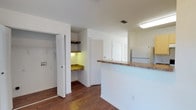 This two-floor residence offers a practical and well-organized layout designed for comfortable everyday living. The kitchen area is a central highlight, featuring a peninsula-style breakfast bar topped with granite-look countertops that provides additional prep space and casual seating. White cabinetry keeps the area feeling bright and clean, while a full-size refrigerator is conveniently positioned nearby. A distinctive built-in nook with matching countertops and open shelving offers a versatile area ideal for use as a home office or study station, complete with under-shelf lighting that adds a warm, functional glow. Adjacent to this area, louvered bifold doors open to a spacious closet fitted with hanging rods and shelving, providing generous storage solutions. Warm hardwood-style flooring runs throughout the space, adding a consistent and inviting aesthetic. The interiors are finished in crisp white walls and ceilings, contributing to a clean, airy ambiance. Recessed ventilation and flush-mounted ceiling lighting ensure the space remains comfortable and well-lit. Spanning two floors, this residence balances practicality with thoughtful design details throughout.