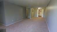 This single-floor residential space offers a clean, functional layout designed for comfortable everyday living. The main living area is expansive and open, finished with plush neutral-toned carpeting that extends throughout, creating a warm and cohesive feel underfoot. The walls are painted in a soft, muted gray tone that lends a calm and modern aesthetic to the interior.

A standout feature of the main room is its generous open floor plan, providing ample space for a variety of furniture arrangements. A floor vent indicates a forced-air climate control system, ensuring year-round comfort. The space flows naturally toward an adjacent area, where a ceiling fan with a light fixture provides both air circulation and overhead lighting.

Beyond the main living area, a closet with a visible rod offers convenient built-in storage. Further into the space, a hallway leads to additional rooms, including what appears to be a utility or laundry area and a separate room accessed through a bright, white-framed doorway. White trim and baseboards throughout provide a crisp contrast to the gray walls, adding a polished, finished look. Overall, the space presents a straightforward and practical interior with a neutral palette well-suited to a range of furnishing styles.