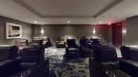 Located at 1600 Harbor Blvd in Weehawken, NJ, this private screening room delivers an upscale, cinema-inspired experience within a single, thoughtfully designed space. The room is furnished with a generous arrangement of premium leather recliner chairs, each equipped with built-in cup holders, offering residents a comfortable and immersive viewing environment. The seating is arranged in a tiered layout facing a wall-mounted flat-screen display, ensuring clear sightlines from every seat in the room. The interior design embraces a dark, sophisticated palette, with textured charcoal and taupe wall coverings that enhance the cinematic atmosphere and minimize light reflection. Recessed ceiling lighting and elegant wall sconces provide subtle, ambient illumination that complements the room's moody aesthetic. The flooring features a bold, abstract-patterned carpet in dark tones with gold and white accents, adding visual interest and contributing to the room's acoustical comfort. A wooden media credenza with glass-paneled cabinet doors anchors one wall, providing a stylish home for audio-visual equipment. The overall ambiance is one of refined luxury, making this screening room an ideal amenity for private movie nights or entertainment gatherings in Weehawken.
