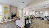 This well-appointed single-floor community space offers a warm and inviting atmosphere designed for comfort and gathering. The centerpiece of the main living area is a striking floor-to-ceiling brick fireplace, flanked by large windows that flood the room with natural light. Soaring vaulted ceilings amplify the sense of openness, while a distinctive patterned carpet ties the generous seating arrangements together. Cream-toned sofas, accent chairs, and dark wood console and coffee tables create an elegant yet approachable aesthetic, complemented by colorful abstract artwork adorning the walls.

The space flows seamlessly into a dedicated dining area, where a wood table and upholstered chairs are positioned alongside a wall of French doors, offering views to the exterior and providing excellent natural illumination. Adjacent to the dining area is a compact kitchenette, featuring light wood cabinetry, a double sink, a full-size refrigerator, a microwave, and granite-style countertops with hardwood-style flooring — well-suited for light catering and hospitality needs.

Throughout the space, neutral wall tones and carefully curated décor contribute to a cohesive, sophisticated ambiance. The layout is practical and versatile, accommodating both intimate gatherings and larger social events with ease.