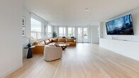 Located at 2453 Renfrew St in Vancouver, BC, this thoughtfully designed single-floor communal space offers a generous, open-plan layout that seamlessly connects multiple functional zones. Light hardwood flooring runs throughout, complementing the bright, white walls and creating a clean, cohesive aesthetic.

The expansive living area is anchored by a large tan leather sectional sofa paired with curved upholstered accent chairs and a distinctive live-edge glass coffee table. Multiple oversized windows flood the space with natural light and frame views of the surrounding Vancouver neighbourhood. Wall-mounted large-screen TVs are positioned throughout for easy viewing from various seating areas.

The kitchen and dining zone is a standout feature, showcasing floor-to-ceiling sage green cabinetry, dark stone countertops, and a substantial island with seating for several. Pendant lighting above the island adds a refined touch, while integrated stainless steel appliances underscore the space's functional design.

A dedicated games area includes a foosball table and wall-mounted display screen, with direct access to an outdoor terrace. A formal dining setup with a dark table and black woven chairs rounds out the space, making it well-suited for both social gatherings and everyday use.