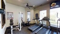 This fitness center offers a practical and comfortable environment for residents to maintain their workout routines. The single-floor space is outfitted with a selection of cardio equipment, including multiple treadmills and a stationary recumbent bike, alongside a cable strength training machine, providing a well-rounded exercise experience. The room features neutral-toned walls and dark carpeted flooring, creating a clean and focused atmosphere. Multiple windows fitted with horizontal blinds line the walls, allowing natural light to filter into the space while maintaining privacy. A ceiling fan mounted centrally overhead aids in air circulation, keeping the environment comfortable during workouts. Recessed lighting supplements the natural light for consistent illumination throughout the room. Informational signage is posted on the wall, likely outlining facility rules and equipment guidelines. The layout is thoughtfully arranged to allow users to move between equipment with ease, making the most of the available space for an efficient and accessible fitness experience.