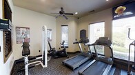 This fitness center offers a practical and comfortable environment for residents to maintain their workout routines. The single-floor space is outfitted with a selection of cardio equipment, including multiple treadmills and a stationary recumbent bike, alongside a cable strength training machine, providing a well-rounded exercise experience. The room features neutral-toned walls and dark carpeted flooring, creating a clean and focused atmosphere. Multiple windows fitted with horizontal blinds line the walls, allowing natural light to filter into the space while maintaining privacy. A ceiling fan mounted centrally overhead aids in air circulation, keeping the environment comfortable during workouts. Recessed lighting supplements the natural light for consistent illumination throughout the room. Informational signage is posted on the wall, likely outlining facility rules and equipment guidelines. The layout is thoughtfully arranged to allow users to move between equipment with ease, making the most of the available space for an efficient and accessible fitness experience.