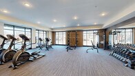 This modern fitness center offers a spacious, single-floor layout designed to accommodate a comprehensive range of workout activities. The facility is thoughtfully organized into distinct functional zones, providing ample room for both cardio and strength training exercises without overcrowding.

The cardio area features multiple treadmills, elliptical machines, and stationary bikes, positioned alongside large floor-to-ceiling windows that invite natural light and offer outdoor views, creating an open and energizing atmosphere. Mounted televisions are available for entertainment during workouts.

The strength training zone is well-appointed with a power rack, cable machine, adjustable benches, and a dedicated free-weight area stocked with a full set of dumbbells. Warm-toned accent walls with wood paneling complement the neutral carpet flooring throughout, contributing to a clean and contemporary aesthetic.

Additional amenities include a water fountain station, storage for fitness accessories such as stability balls, and a dedicated entry area. Recessed ceiling lighting ensures consistent illumination across the entire space. The interior design balances functionality with a polished, modern finish, making this fitness center a well-rounded facility suited for a variety of fitness routines and training styles.