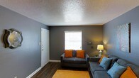 This single-floor space offers a thoughtfully arranged living area that combines comfort and functionality. The living room serves as the heart of the space, finished with cool gray walls that create a modern, neutral backdrop. Dark hardwood flooring runs throughout, providing a warm contrast to the lighter wall tones and adding a sense of depth and richness underfoot. A textured popcorn ceiling adds a layer of visual interest and dimension to the room.

Natural light filters in through a window fitted with white horizontal blinds, casting a bright and inviting glow across the space. The room comfortably accommodates multiple seating areas, with soft gray sofas accented by vibrant orange and blue throw pillows that introduce pops of color and a relaxed, lived-in warmth. A warm floor lamp in the corner contributes to the ambient lighting, creating a cozy atmosphere during evening hours.

A white interior door is visible along one wall, contributing to the clean, simple aesthetic of the space. White baseboards frame the room neatly, offering a crisp finish that ties together the overall design. The layout is efficient and open, making the space feel welcoming and easy to navigate. Overall, this space presents a comfortable and stylish single-level living environment.