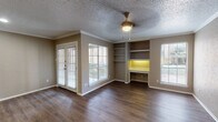 This single-floor unit offers a well-organized layout with distinct living, kitchen, bedroom, and bathroom areas. The living room serves as the centerpiece of the space, showcasing rich hardwood-style flooring, crown molding, and a textured ceiling with a central ceiling fan. A standout feature is the built-in shelving and illuminated desk nook along one wall, providing both functional workspace and display storage. French doors and large windows fitted with horizontal blinds allow generous natural light while offering outdoor access.

The kitchen is accessible via a pass-through opening from the main living area, creating an open, connected feel. It features white cabinetry, light quartz-style countertops, a bold black tile backsplash, and a full suite of black appliances including a refrigerator, electric range, and over-the-range microwave.

The bedroom is a comfortable, carpeted room with multiple windows and a ceiling fan, creating a bright and restful environment. The bathroom includes a white vanity with ample counter space, a large mirror with overhead lighting, and a shower-tub combination. Hardwood-style flooring continues into the bathroom, maintaining a cohesive aesthetic throughout the unit.