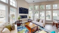 Located at 1314 Kingston Way in Chesapeake, VA, this single-floor space offers an open-concept layout that seamlessly connects the living room, dining area, and kitchen. Soaring vaulted ceilings amplify the sense of space throughout, while light wood-tone flooring flows consistently across all areas.

The living room serves as the focal point of the space, featuring a sleek linear fireplace, a wall-mounted flat-screen television, and a ceiling fan. Large windows on multiple walls flood the room with natural light and frame views of the surrounding greenery outdoors. A vibrant, multi-colored area rug adds a bold design element to the neutral, warm-toned interior.

The dining area is defined by an expansive wall of tall windows that further maximizes natural light and outdoor views. High-top dining tables with barstool seating create a casual yet functional atmosphere well-suited for gatherings.

The kitchen is designed for both style and efficiency, showcasing white shaker cabinetry extending to the tall ceilings, granite countertops, and stainless steel appliances including a refrigerator and built-in microwave. A granite-topped island with bar seating provides additional prep space and serves as a natural gathering point, completing this cohesive and well-appointed space.