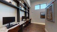 This single-floor space is thoughtfully designed to blend functionality with modern aesthetics. The layout includes two distinct areas: a built-in workstation zone and a mailroom corridor, each exhibiting clean architectural lines and carefully selected finishes.

The workstation area features an extended granite-topped desk running along a dark navy built-in shelving unit, equipped with recessed lighting that illuminates displayed items and provides focused task lighting. Multiple computer workstations are arranged along the desk, accompanied by ergonomic leather chairs. Soaring ceiling height draws the eye upward, and a clerestory window near the top of the wall introduces natural light without sacrificing wall space. A cork bulletin board is mounted nearby, adding a practical organizational element. Wood-look tile flooring runs throughout, providing a warm, cohesive foundation.

The mailroom corridor presents rows of dark bronze individual mailboxes lining both walls, accented by warm LED underlighting at the base that creates a welcoming glow along the floor. Gold tubular pendant lights hang from the elevated ceiling, lending an upscale touch to the otherwise utilitarian space. The corridor connects visibly to the workstation area, reinforcing the open, interconnected flow of the overall layout.